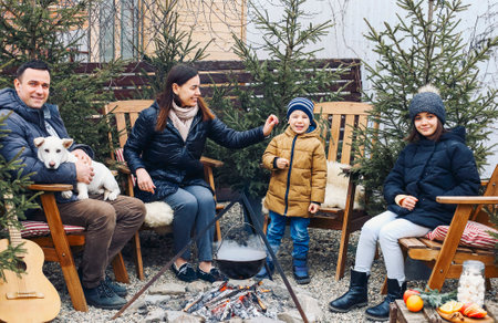 New Years gatherings by winter bonfire. Cozy portrait of happy family parents, two children and sweet dog cooking food in steaming pot over campfire while sitting in backyard outdoorsの写真素材