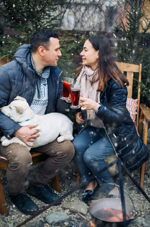 Lovely couple with cute dog sitting on backyard outside surrounded by young coniferous trees,...