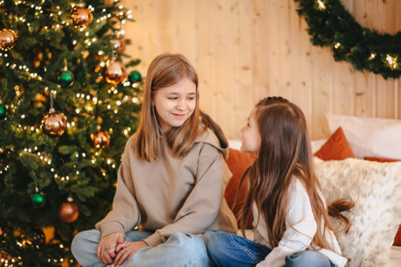 Cozy domestic portrait of two happy smiling little girls sisters hugging embracing while spending leisure time together at home, sitting on bed near decorated Christmas tree. Family and children conceptの写真素材