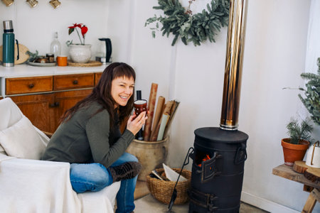 Back view photo of woman sitting near potbelly stove at home, female warming hands after winter walk in frosty weather, New Years home decor around. People and winter holidays conceptの写真素材