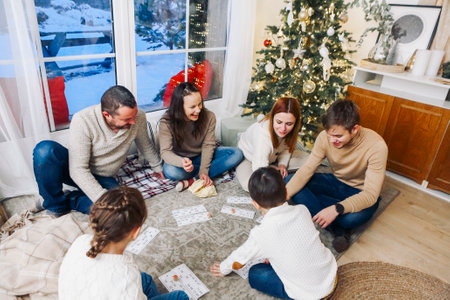 Big family with children sitting on floor near Xmas tree playing lotto board game together while spending leisure time at cozy home during Christmas holidays. Indoor winter activitiesの写真素材