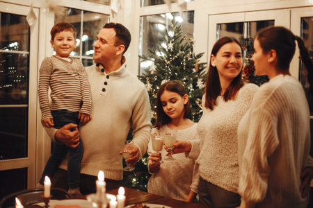 Happy joyful family parents with two kids pouring homemade eggnog into glasses while standing behind wooden table together and cooking in cozy rustic kitchen during winter holidaysの写真素材