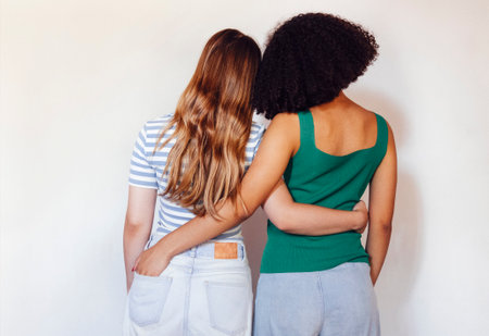 Strong female friendship. Rear view two teen girls best friends holding hands behind back and hugging while standing in front of beige wall outdoorsの写真素材