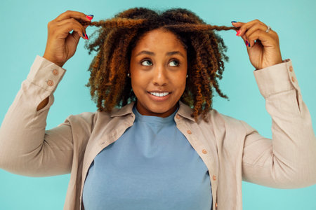 Positive african american female model smiling at camera and touching clean curly hair after hygiene...
