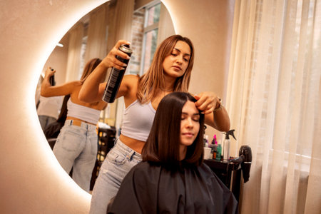 Hairstyling process in beauty salon. Female hairdresser fixing customer woman hair with spray and put care treatment while styling hairdress for client. beauty and fashion serviceの写真素材