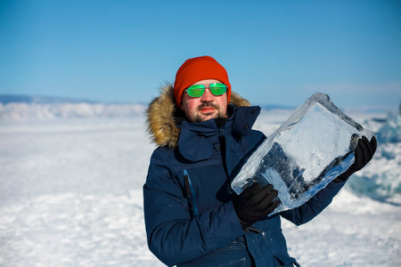 Happy smiling man wearing winter blue jacket and red hat holding piece of transparent crushed ice...