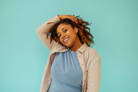 Positive african american female model smiling at camera and touching clean curly hair after hygiene treatment on blue backgroundの写真素材
