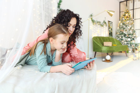 Family Christmas morning. Beautiful family mother and little daughter in pajamas watching movie or cartoon together on modern digital tablet while relaxing on canopy bed decorated with xmas lightsの写真素材