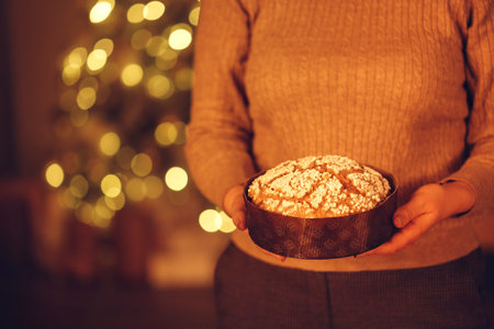 Cropped shot of woman holding delicious freshly baked homemade Christmas pie while standing against blurred xmas tree in background, preparing food for New Years Eve, selective focus on panettone cake in handsの写真素材