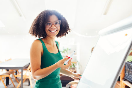 Beautiful African American girl with dark curly hair standing near board and giving presentation to colleagues in office. Young business woman looking on her coworkers while discussing new projectの写真素材