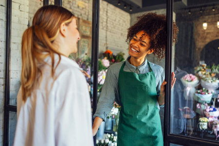 Happy black woman entrepreneur standing in plant store selling fresh flowers to client. Young blond girl buy a fresh bouquet from florist. Smiling african woman botanist, selling flowersの写真素材