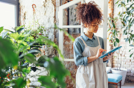 Young black woman wearing beige apron on flower shop background with copy space. Portrait of successful african american woman looking at tablet computerの写真素材