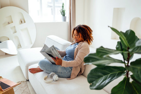Photo of pretty happy young african woman sitting on sofa indoors at home while reading magazine or bookの写真素材