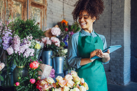 Young black woman wearing beige apron on flower shop background with copy space. Portrait of successful african american woman looking to the flowers and holding tablet computerの写真素材