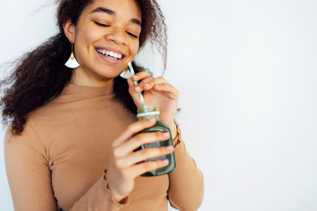 Fitness afro woman holding a vegetable drink. Young african american female drinking green juice with reusable bamboo straw in loft apartment. home concept. Healthy lifestyle concept. copy spaceの写真素材