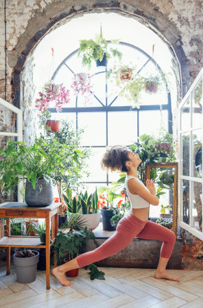 Young adult happy fit slim healthy African American ethnic woman wearing sportswear stretching in yoga pose at home by the window, doing fitness morning workout exercises training.の写真素材