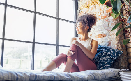 Pleased relaxed African American ethnicity girl in homewear sitting on windowsill, happy afro female at home smiling gently looking to the windowの写真素材