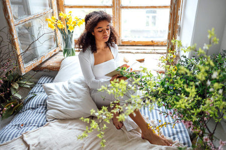 Pretty romantic african american girl, reading book sitting on windowsill, suraunded with flowers. Beautiful Afro woman happily reading a novel in cozy room. education conceptの写真素材