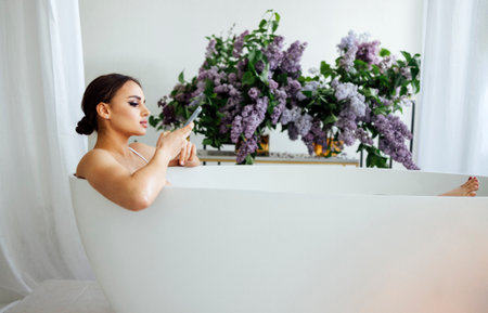 Side view portrait of a pretty asian woman typing message on mobile phone while having a bath at homeの写真素材