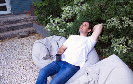 Brunette handsome man freelancer sitting on chair holding cup of coffee at front yard outside at...