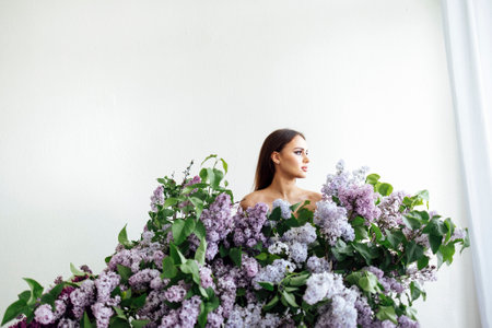 Profile portrait of a happy young woman in a white shirt by bouquets of lilacs all arounded her like dress. Beautiful smiling white brunette female in profile on a light background. studio portrait.の写真素材