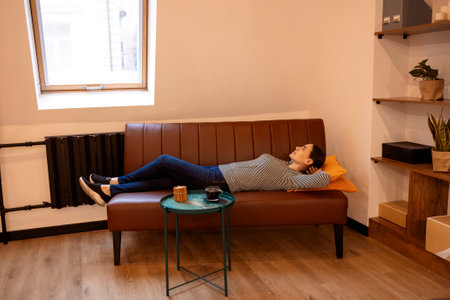 Serene housewife lady, living room, sitting on sofa, feeling tired, holding hands behind head, calm young woman resting on comfortable sofa, breathing fresh air in cozy clean modern living room at homeの写真素材