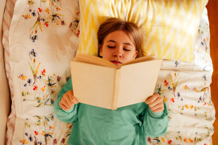 Caucasian blond teenage girl is lying on the bed and reading book. Reading fiction, hobbies, leisure...