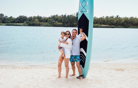 Joyful married couple of surfers with their cute daughter on the beach. Smiling man holding paddleboard and hugging woman in sunglasses, swimsuit and white shirt holding little girl in her arms.の写真素材