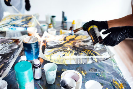 A woman uses a gas burner to draw in Fluid Art. Process of creating a painting with liquid acrylic paints. Paintings, paints, brushes and plastic glasses on the table. close up. Acrylic Fluid Pouring.の写真素材