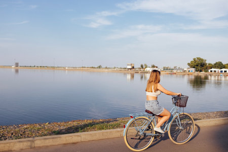 Back view of female teenage is driving her bicycle. Young and positive girl in casual clothes has a fun. City street and trees with bushes in the background. copyspace.の写真素材