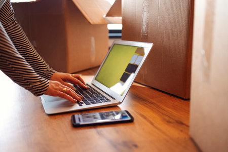 Female hands are typing on a laptop on the parquet floor or table. There are a telephone nearby and a lot of cardboard boxes with things. The concept of moving from an apartment or house. copyspace.の写真素材
