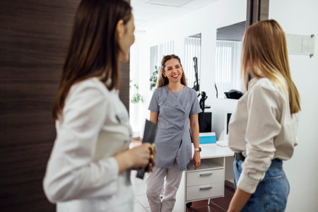 Joyful female beautician meets her new client. Three women communicate and smile at each other. Welcoming young girl in casual clothes in beauty salon. Light modern interior in clinic. copyspace.の写真素材