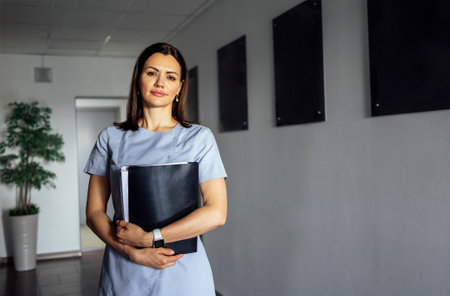 Beautiful smiling female doctor in medical uniform stands near the window in clinic. Young nurse is holding folder of paper in her hands. Sunny day. light interior. copyspace.の写真素材