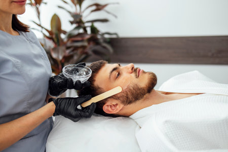 Beauty salon beautician applies cooling gel to clients skin. Preparing the skin of young man for laser hair removal of the cheek. Close up of brunette male in clinicの写真素材