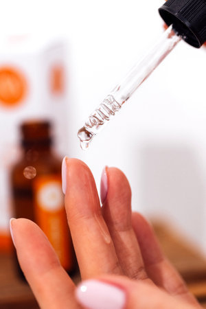 Extreme close up of woman's hand and pipette. Transperent drop of essence, oil or gel drips from the pipette. Blurred brown glass serum bottle on the background. Concept of natural organic cosmeticsの写真素材