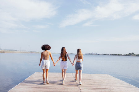 Three teens girls of different nationalities joined hands and walk along the wooden bridge near a river or lake. view from the back. Positive teenagers have a fun together. friendship concept.の写真素材