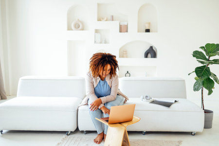Young African American woman is working on a laptop. Happy female student is looking at computer screen and watching webinar or doing video chat by webcam. remote education or home office conceptの写真素材