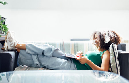 Pretty smiling african american woman is lying on the sofa with a laptop and headphones. Happy young woman enjoying music or listening to a webinar. Dark skin female is freelancer or studentの写真素材