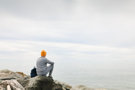 Young man in casual clothes sits on the rocks near the sea. Caucasian guy in a blue sweater and hat holds his backpack and looking at the calm sea. view from the back. The concept of nature and peaceの写真素材