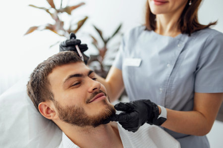 beauty injections. Female cosmetologist in transparent gloves makes an injection in the lower jaw of her client. Treatment with hyaluronic acid. Introduction of liquid into the skin of young manの写真素材