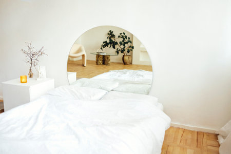 Double bed with white linens and square nightstand. At head of bed is a large round mirror. Reflection shows small coffee table, armchair and flower pot. Light stylish bedroom interior.の写真素材