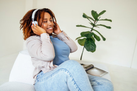 Body positive curvy afro american woman in headphones on a light sofa at home. Young dark skinned woman in casual clothes closed her eyes and enjoys the music. Light interior on the background.の写真素材