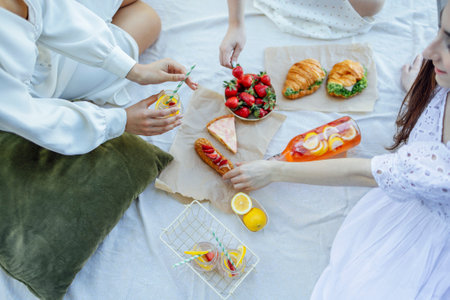 Happy female friends on picnic on the bank of a river. Blanket with pillows and food on a wooden bridge. Bottle of strawberry and lemon smoothie, soda glasses with straws, herb croissants, sweet cakes and fresh strawberriesの写真素材