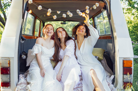 Pretty smiling three teenage girls in light dresses. Female teens of different nationalities sit at retro minibus. Old car is decorated with macrame panel. Boho style. copyspace.の写真素材