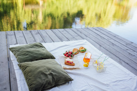Picnic on the bank of a river. Blanket with pillows and food on a wooden bridge. Bottle of strawberry and lemon smoothie, soda glasses with straws, herb croissants, sweet cakes and fresh strawberriesの写真素材