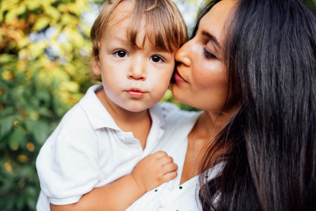 Lovely portrait of a beautiful mother kissing her little son. Close up of a young brunette female face touching her cute child. green leaves in the background. copy space.の写真素材