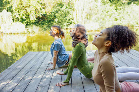 Group of teenages is engaged in fitness on pier by the lake or river. Young and positive girls go in for sports. Muslim in a hijab, african american and white females are doing stretching in the park.の写真素材