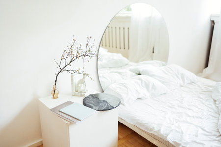 Double bed with white linens and square nightstand. At head of bed is a large round mirror. Reflection shows small coffee table, armchair and flower pot. Light stylish bedroom interior.の写真素材