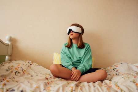 Pretty female teenager is sitting in VR glasses on bed in her bedroom. Blonde girl in relaxed pose looks up. Modern light room interior. Free space for text.の写真素材