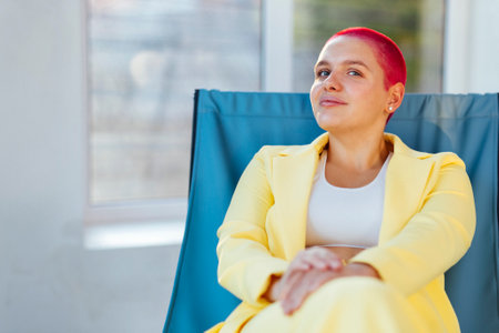 Beautiful relaxed woman in yellow suit and red shot hair sitting in blue armchair while resting on sunny weekend day at home. concept of body positiveの写真素材